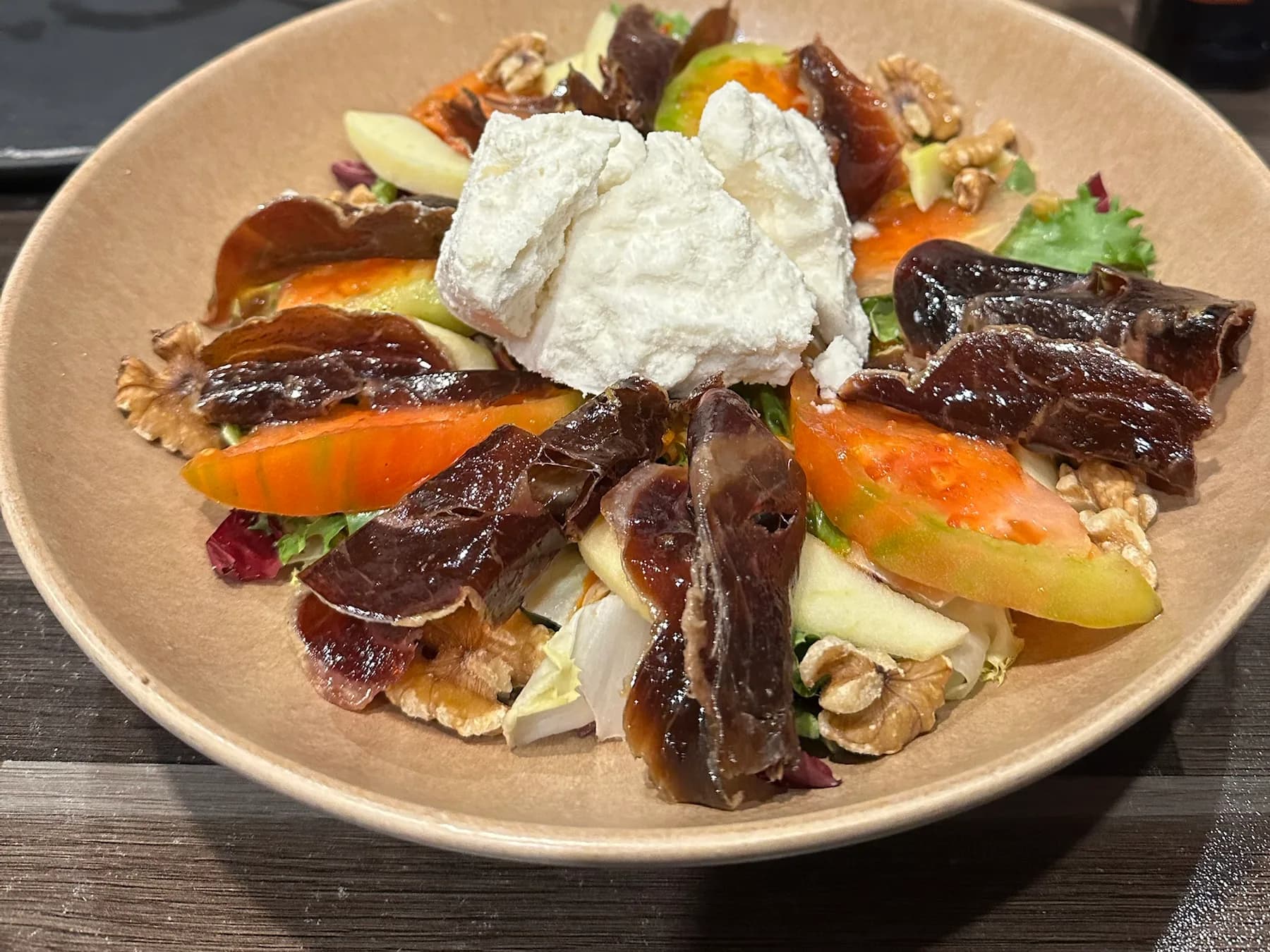 Ensalada de cecina de Tixola Tapería: láminas de cecina repartidas en corona sobre hojas de lechuga y endivia, con gajos de tomate, láminas de manzana y nueces, y una quenelle de helado de queso de oveja en el centro, en un cuenco hondo de cerámica sobre la mesa de madera